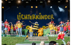 Download Foto: Kinder auf einer Bühne in bunten Kostümen, mit dem Schriftzug 'Lichterkinder' im Hintergrund. Fröhliche Atmosphäre.