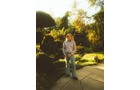 Download Foto: Eine Person steht in einem sonnigen Garten auf einem gepflasterten Weg, umgeben von Bäumen und Sträuchern.