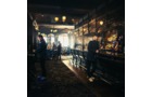 Download Foto: Eine stimmungsvolle Bar mit dunkler Inneneinrichtung, mehreren Personen an der Theke und warmem Licht, das durch große Fenster fällt.