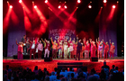 Download Foto: Eine Gruppe von Menschen steht auf einer Bühne unter roten Lichtern, alle heben eine Hand in die Luft. Im Hintergrund 'Queer Vocal' Schriftzug.