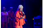 Download Foto: Ältere Person in rotem Outfit, singt auf Bühne mit Mikrofon, blaue Beleuchtung, zwei Personen im Hintergrund.