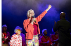 Download Foto: Eine Person mit Mikrofon singt auf einer Bühne, hebt die Faust; umgeben von anderen Personen in bunter oder roter Kleidung.