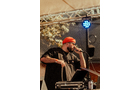 Download Foto: Ein DJ mit roter Mütze performt auf einer Bühne, hält ein Mikrofon und steht vor DJ-Equipment unter einem beleuchteten Pavillondach.