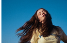 Download Foto: Eine Frau mit langen Haaren genießt mit geschlossenen Augen die Sonne vor klarem, blauem Himmel. Sie trägt ein helles T-Shirt.