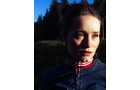 Download Foto: Eine Frau steht im Sonnenschein, trägt eine blaue Jacke und einen roten Schal. Im Hintergrund sind Bäume und blauer Himmel zu sehen.