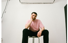 Download Foto: Ein Mann in einem rosa T-Shirt sitzt entspannt auf einem Hocker in einem weißen Raum, die Hand am Kinn.