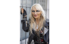 Download Foto: Person mit langem blondem Haar, schwarzer Lederjacke und Handschuhen lehnt lächelnd an einem Metallgitter.