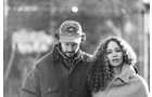 Download Foto: Ein Schwarz-Weiß-Foto zeigt einen Mann mit Mütze und eine Frau mit lockigem Haar, nebeneinander draußen stehend.