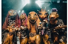 Download Foto: Fünf Dinosaurier in bunten Kostümen und mit Instrumenten posieren auf einer Bühne mit hellem Scheinwerferlicht im Hintergrund.