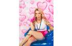 Download Foto: Frau mit Tattoos sitzt auf einem blauen Stuhl vor rosa Herz-Hintergrund, trägt ein weißes Top und pinke Shorts.