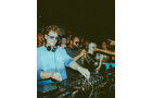 Download Foto: Ein DJ trägt eine Sonnenbrille und mischt an einem Pult in einem Club, umgeben von feiernden Menschen im gedämpften Licht.