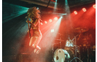 Download Foto: Musiker springt mit E-Gitarre auf der Bühne, umgeben von bunten Lichtern und einem Schlagzeug im Hintergrund. Atmosphäre ist energisch.