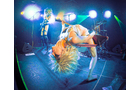 Download Foto: Ein Performer mit blonden Haaren tanzt rückwärts auf einer Bühne mit bunten Scheinwerfern im Hintergrund.