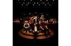 Download Foto: Eine Gruppe von sechs Personen sitzt und steht auf einer Bühne in einem warm beleuchteten Theaterambiente, umgeben von leuchtenden Lichtern.