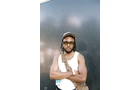 Download Foto: Eine Person mit Sonnenbrille und Mütze steht mit verschränkten Armen vor einer dunklen Wand. Sie trägt ein weißes Tanktop.
