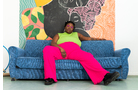 Download Foto: Person in grüner Bluse und pinker Hose sitzt entspannt auf blauem Sofa vor einer bunten Wand mit abstrakten Motiven.