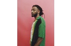 Download Foto: Ein Mann mit Dreadlocks trägt eine grüne und gelbe Musterjacke. Er steht vor einem pinkfarbenen Hintergrund und schaut zur Seite.