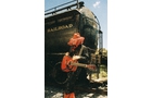 Download Foto: Ein Mensch mit Cowboy-Hut spielt Gitarre vor einem alten Eisenbahnwaggon im Freien.