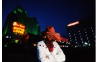 Download Foto: Ein Cowboy in weißer Jacke steht vor beleuchteten Gebäuden bei Nacht, darunter ein Neon-Schild mit der Aufschrift 'Monte Carlo'.