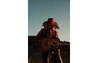 Download Foto: Eine Person mit Cowboyhut spielt Gitarre in einer Landschaft bei Sonnenuntergang, der Himmel ist klar.