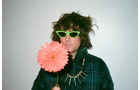 Download Foto: Person mit grüner Sonnenbrille, zerzaustem Haar, trägt blauen Mantel, hält große pinke Blume und trägt auffällige Halskette.