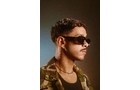 Download Foto: Ein Mann mit Sonnenbrille und lockigem Haar trägt eine Camouflage-Jacke, im Profil vor einem dezent beleuchteten Hintergrund.