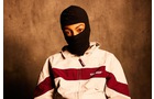 Download Foto: Eine Person in einem schwarz-weißen Kapuzenpullover mit rot-weißem Streifen trägt eine schwarze Skimaske, vor braunem Hintergrund.