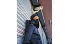 Download Foto: Eine Person mit Sturmhaube lehnt an einem geschlossenen Rolltor und schaut zu einer Überwachungskamera hinauf.