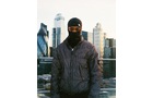 Download Foto: Eine Person in einer dunklen Jacke und Sturmhaube steht vor einer städtischen Skyline mit modernen Gebäuden im Hintergrund.