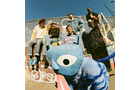 Download Foto: Eine Gruppe von fünf Personen sitzt auf einer Rampe, im Vordergrund ein großes, blaues Maskottchen mit großen Augen und Zunge.