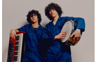 Download Foto: Zwei Personen in blauen Overalls stehen nebeneinander. Eine hält ein Keyboard, die andere eine Trommel.