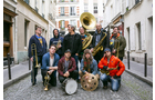 Download Foto: Eine Gruppe von zwölf Musikern mit verschiedenen Blasinstrumenten und einer Trommel posiert in einer gepflasterten Gasse.