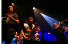 Download Foto: Eine Band auf der Bühne spielt Blasinstrumente unter bunten Lichtern, Musiker sind im energetischen Spiel vertieft.