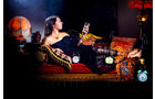 Download Foto: Frau in schwarzem Kleid liegt auf einem orientalisch dekorierten Sofa, umgeben von Uhren und einem Schachbrett.