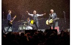 Download Foto: Eine Band spielt vor einem großen Publikum im Regen. Ein Sänger hat die Arme ausgebreitet, während Gitarristen und ein Schlagzeuger spielen.