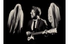 Download Foto: Ein Mann im Anzug spielt E-Gitarre, umgeben von zwei geisterhaften Gestalten in Laken, vor einem schwarzen Hintergrund.