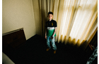 Download Foto: Ein Junge steht in einem Zimmer vor einem großen Fenster mit zugezogenen Vorhängen, in legerer Kleidung mit Jeans und dunklem Shirt.