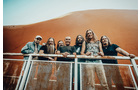 Download Foto: Gruppe von sieben Personen steht lächelnd auf einem Geländer. Im Hintergrund ist eine rote Sanddüne zu sehen.