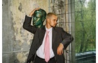 Download Foto: Person in Anzug und rosa Krawatte lehnt an einer beschädigten Wand und hält eine grüne Büste auf dem Kopf. Draußen sind Bäume sichtbar.
