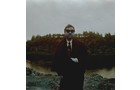 Download Foto: Ein Mann in dunklem Anzug und Sonnenbrille steht draußen vor bewölktem Himmel, Wald im Hintergrund.