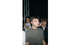 Download Foto: Eine Person mit kurzem Haar steht auf einer Veranstaltung, umgeben von anderen Menschen. Hintergrund: dunkle Beleuchtung und Bühnenträger.