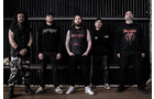 Download Foto: Fünf Männer stehen nebeneinander vor einer dunklen Wand, alle in schwarzen Band-Shirts und Hosen gekleidet, mit ernstem Gesichtsausdruck.