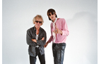 Download Foto: Zwei Personen stehen vor weißem Hintergrund, eine trägt Sonnenbrille und eine pinke Jacke, die andere eine dunkle Jacke.