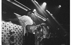 Download Foto: Schwarz-weiß-Bild einer Band auf der Bühne, Gitarrist im Vordergrund mit ausgestreckten Armen, Scheinwerfer beleuchten die Szene.