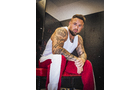 Download Foto: Ein Mann mit Tattoos sitzt auf einem Stuhl, trägt ein weißes Tanktop und rote Hosen. Er schaut direkt in die Kamera.