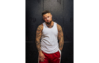 Download Foto: Mann mit Tattooärmeln trägt weißes Tanktop und rote Hose, steht vor dunkler Wand und schaut direkt in die Kamera.