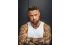 Download Foto: Ein tätowierter Mann in einem weißen Tanktop steht mit verschränkten Armen vor einem dunklen Hintergrund.