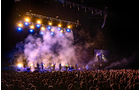 Download Foto: Bühnenansicht eines Konzerts bei Nacht, mit bunten Lichtern, Rauch und einer großen Menschenmenge davor.