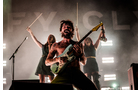 Download Foto: Ein Musiker spielt leidenschaftlich Gitarre auf der Bühne, während zwei Personen im Hintergrund ihre Arme mit Drumsticks heben.