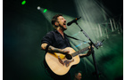 Download Foto: Ein Musiker singt leidenschaftlich in ein Mikrofon und spielt dabei eine Akustikgitarre auf einer Bühne mit grüner Beleuchtung.